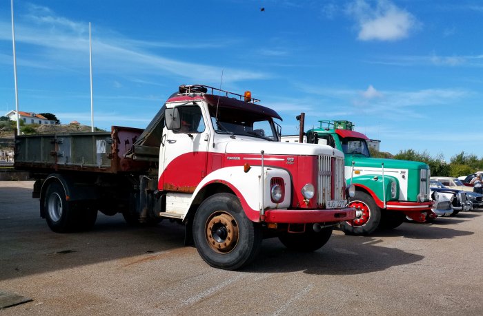 Vintage röd och vit Scania lastbil parkerad framför en grön och vit Scania på en solig parkeringsplats.