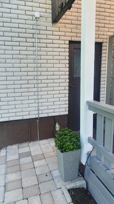 Kabelskyddsrör längs en husvägg med en ansluten eldosa och plantering vid husets entré.