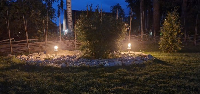 Trädgårdsbelysning i natt med upplysta lampor runt en buske, stenar och ett staket i bakgrunden.