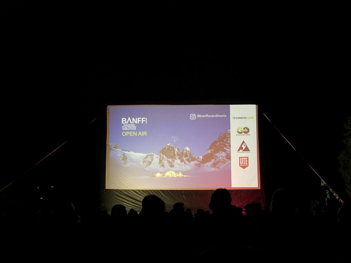 Utomhusbiograf med publik som ser på en skärm visande 'BANFF Mountain Film Festival OPEN AIR' med berg i bakgrunden.