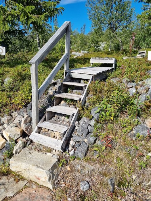 Trätrappa med räcke utomhus i en stenig trädgård, omgivet av grönska och träd under sommarhimlen.