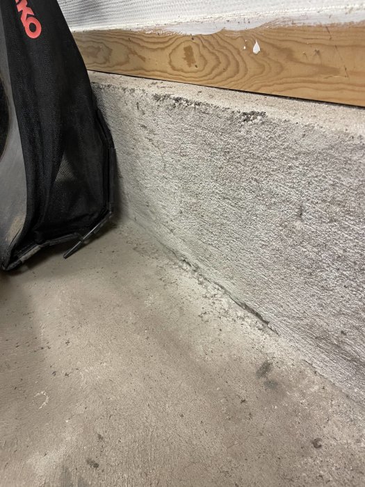 Hörn där betonggolv möter betongvägg med synliga fuktskador och svart tygdel från en väska.