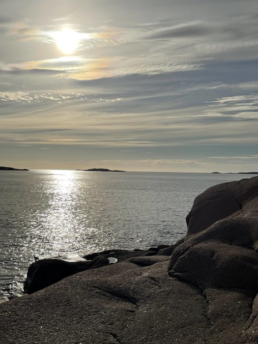 Bild av klippor vid havet med solljus som reflekteras på vattenytan och moln på himlen.