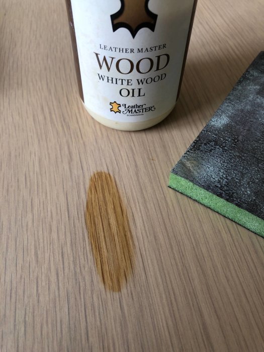 Flaska av "Leather Master Wood White Wood Oil" bredvid en träyta med olja applicerad och en slipkloss.