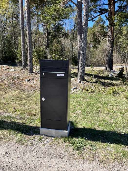 Svart paketlåda på gjuten sockel i gräsmiljö med skog och grusväg i bakgrunden.