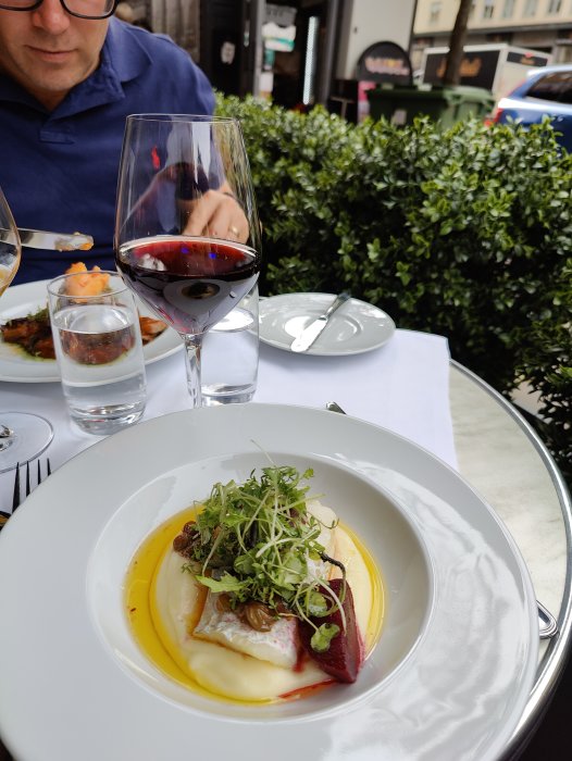 Elegangt upplagd maträtt på restaurang med ett glas rödvin och en person i bakgrunden.