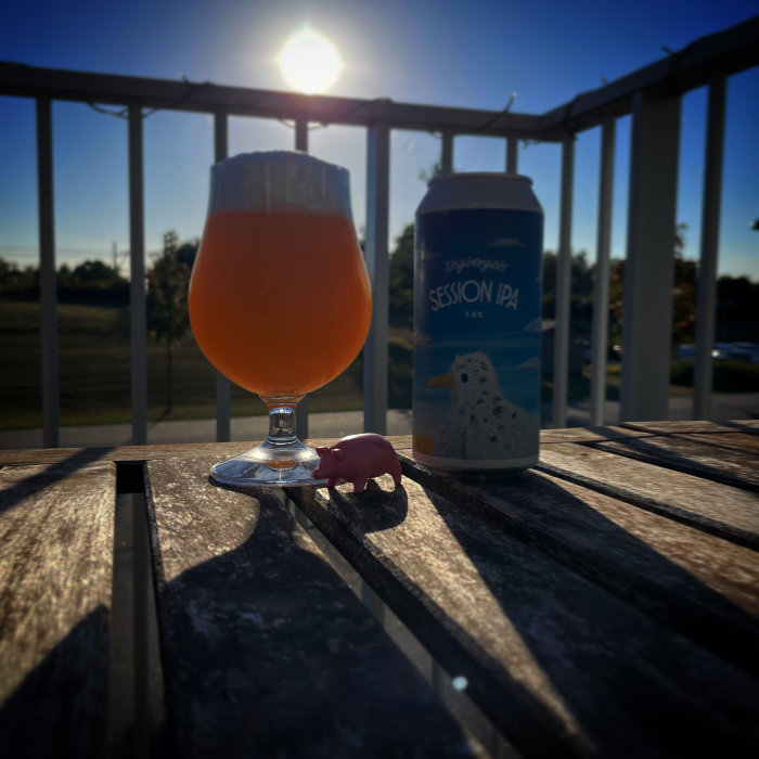 Ett glas öl med skumkrona på ett bord vid sidan av en burk Session IPA, mot solljus och en liten rosa plastgris.