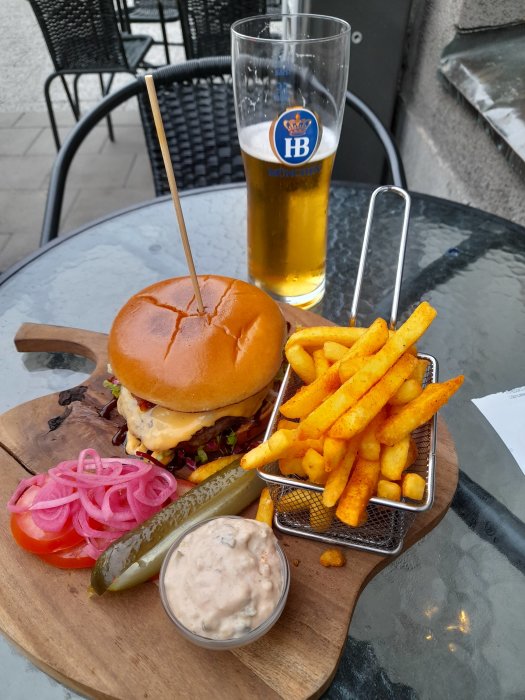 En hamburgare med tillbehör och ett glas öl på ett utomhusbord på restaurang.