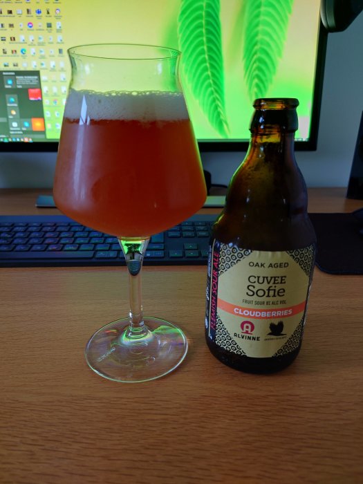 Ett glas med hjortronsuris bredvid en ölflaska, märkt Cuvee Sofie Cloudberry, på ett skrivbord.