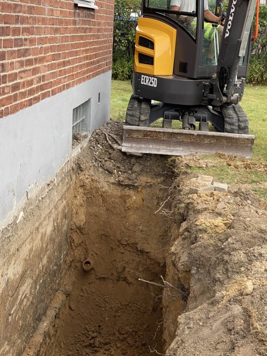 Minigrävare intill en husgrund gräver en djup grop i en trädgård.