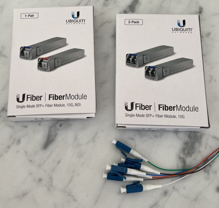 UBIQUITI fibermoduler och optiska kablar på marmor yta, 1- och 2-pack lådor visar 10G SFP+ produkter.