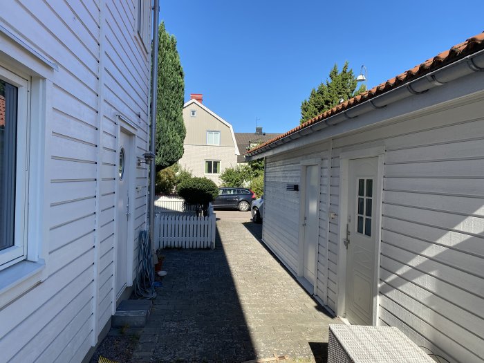 Trång uppfart mellan vitmålade hus med staket och garage på en solig dag.