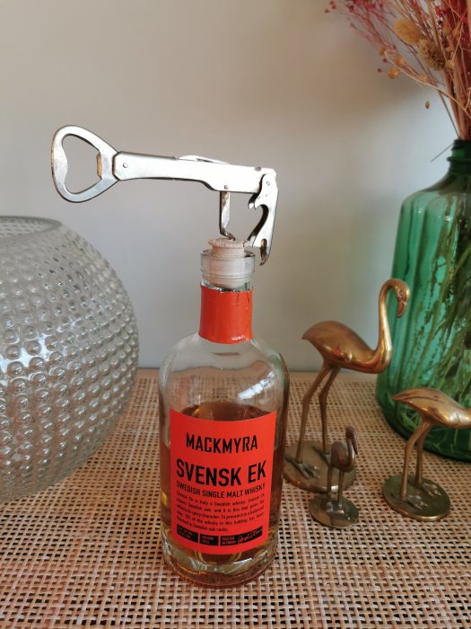 Flaska av Mackmyra Svensk Ek whisky med öppnare på toppen mot en bakgrund av en vas och figuriner.