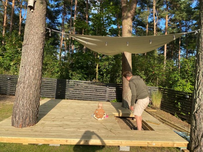 Man bygger ett trädäck runt träd, med en bäbis som tittar på och en hängmatta i bakgrunden.