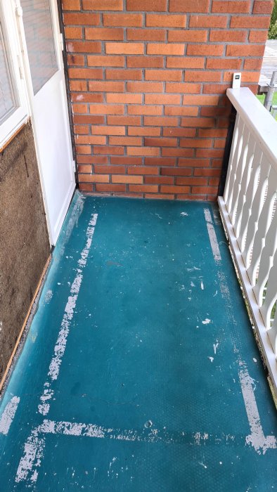 Utsliten blå balkongmatta på betonggolv med vit dörr och tegelvägg till vänster, vit räcke till höger.