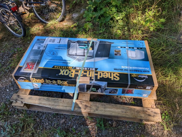 Förpackad förrådstält 'Shed-in-a-Box' på en träpall med cykel i bakgrunden.