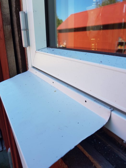 Närbild på en smutsig vit fönsterbleck vid ett stängt fönster med reflektioner och röd byggnad i bakgrunden.