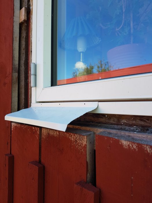 Fönster med vit fönsterbleck monterad på rödmålad vägg, reflektion av taklampa i glaset.