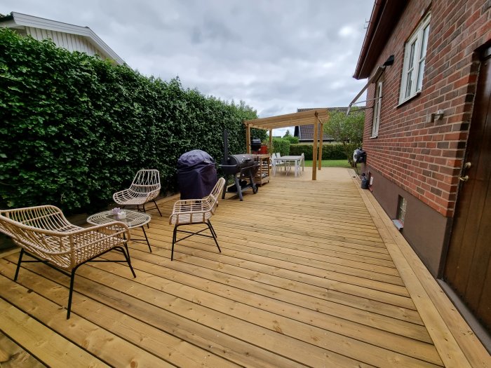 Nybyggd trädäck med rottingmöbler, grill och pergola bredvid en tegelbyggnad och häck.