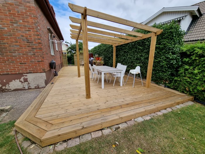 Trädäck med pergola nära tegelhus, vit utemöbler och halvrenoverad grundmur syns.