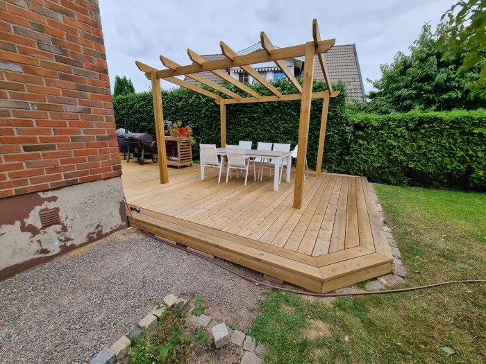 Nybyggd trädäck med pergola, vit trädgårdsmöbel, på halvfärdig grundmur vid tegelhus.