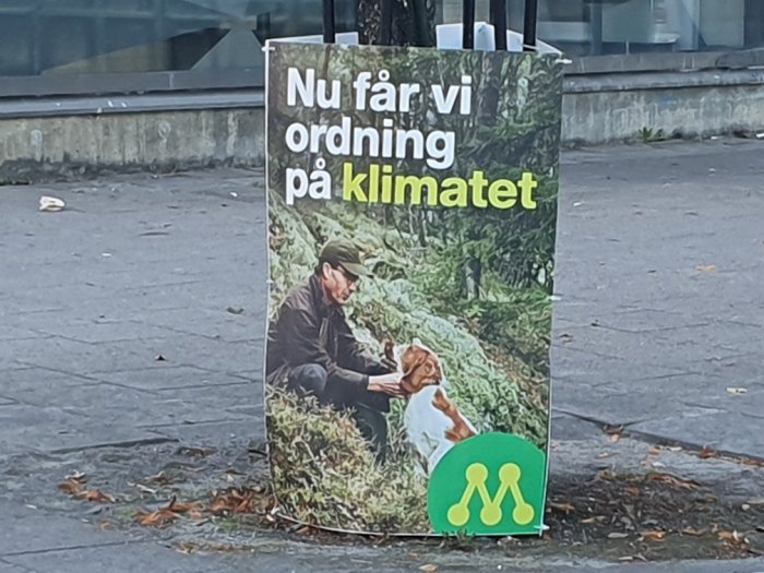 Reklamskylt utomhus med texten "Nu får vi ordning på klimatet" och bild av person med spade och hund.