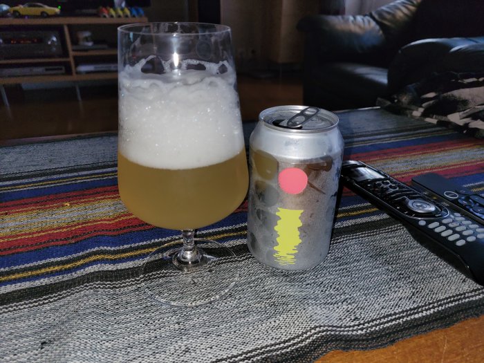 Öppen burk och glas med fruktig IPA på ett färgglatt dukat bord, fjärrkontroll i bakgrunden.