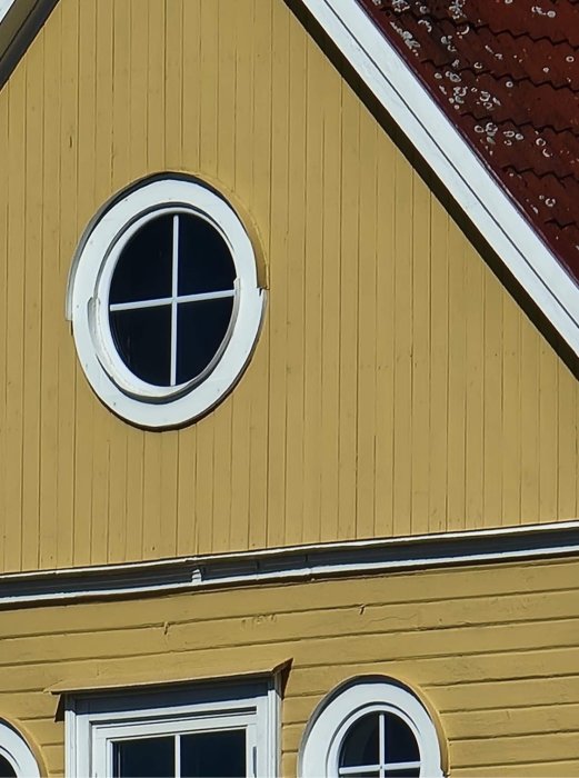 Runt ovanvåningsfönster på gul panelvägg med del av tak och fönster på nedre våningen.