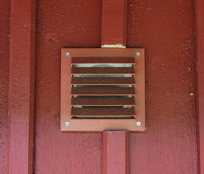 Röd ventilationsgaller monterad på en röd trävägg på ett fritidshus.