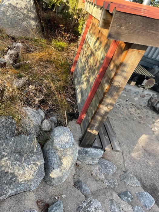 Stolpstöd vid trädäck som vilar på sten på en ojämn terräng med gräs och små buskar.