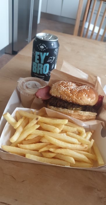 Hamburgare och pommes frites i take-away förpackning med en läskburk märkt "HEY BRO" på ett träbord.