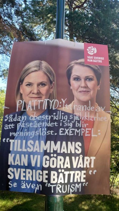 Affisch på lyktstolpe med politiska budskap och två kvinnoporträtt i förgrunden, omgiven av träd.