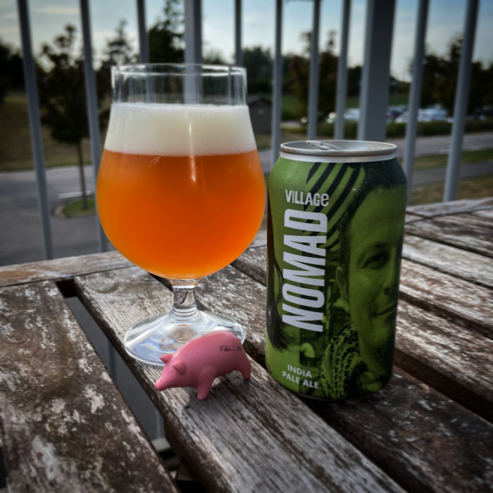 Ölglas med orangefärgad dryck bredvid en ölburk och en liten rosa spargris på ett träbord utomhus.