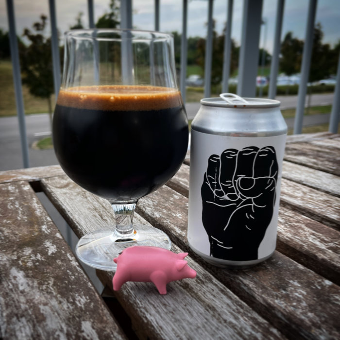 Ölglas med mörkt skummande öl och ölburk med handmotivbredvid en rosa plastgris på ett träbord utomhus.