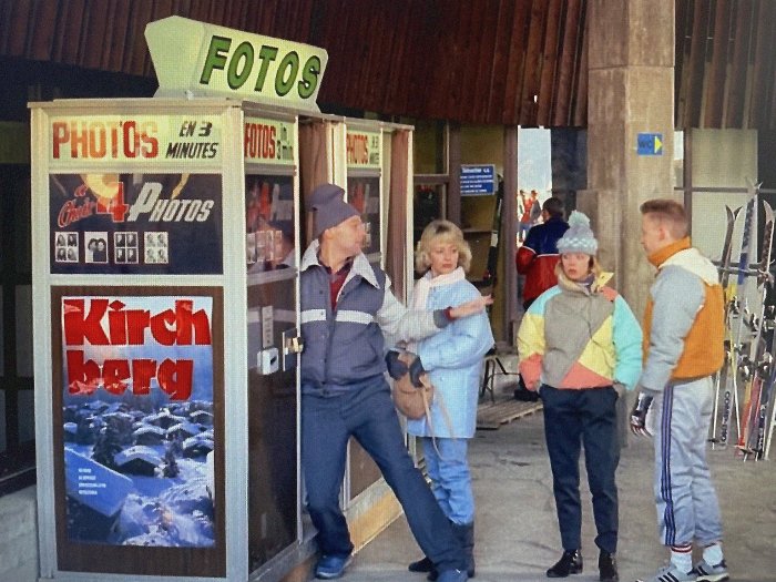 Människor i vinterkläder utanför en fotoautomat med skylten "FOTOS" och en reklamposter för "Kirchberg".