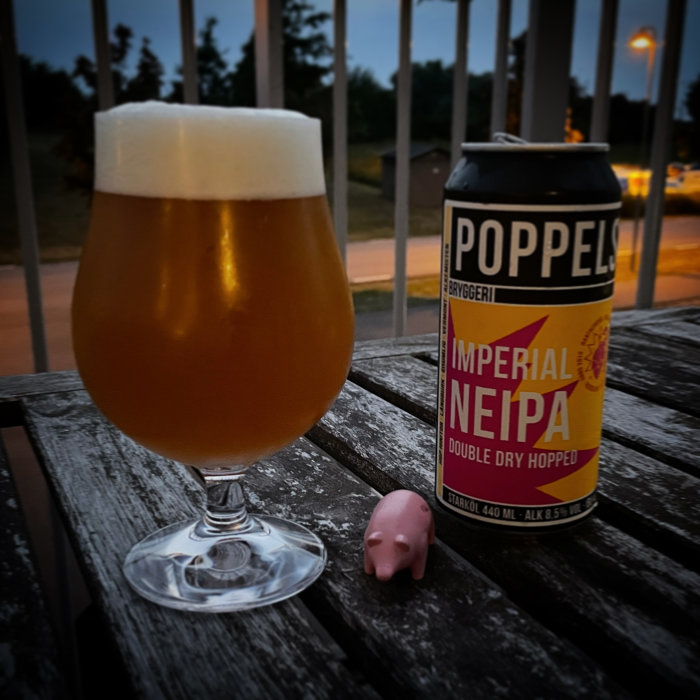 Ölglas med skum på träbord bredvid en ölburk från Poppels och en liten rosa plastgris.