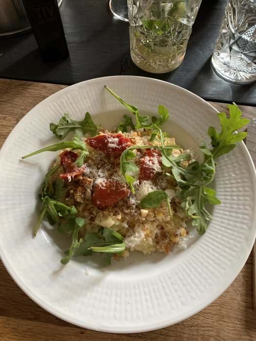 Tallrik med majsrisotto toppad med riven ost, soltorkade tomater och ruccola på ett mörkt bord.