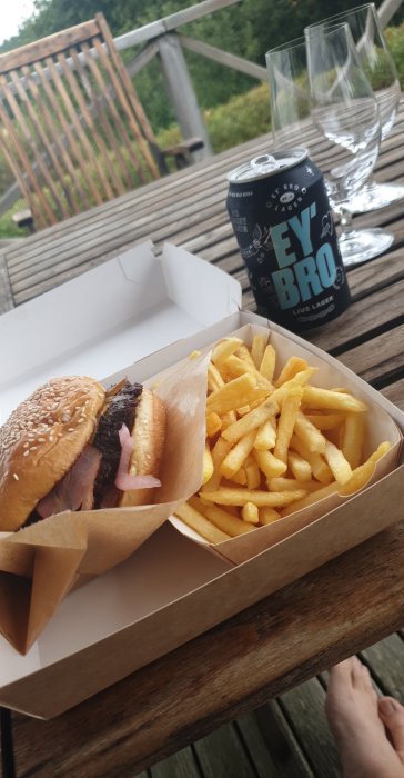 Hamburgare och pommes frites i take-away förpackningar på ett träbord utomhus med en burk öl och tomma glas.