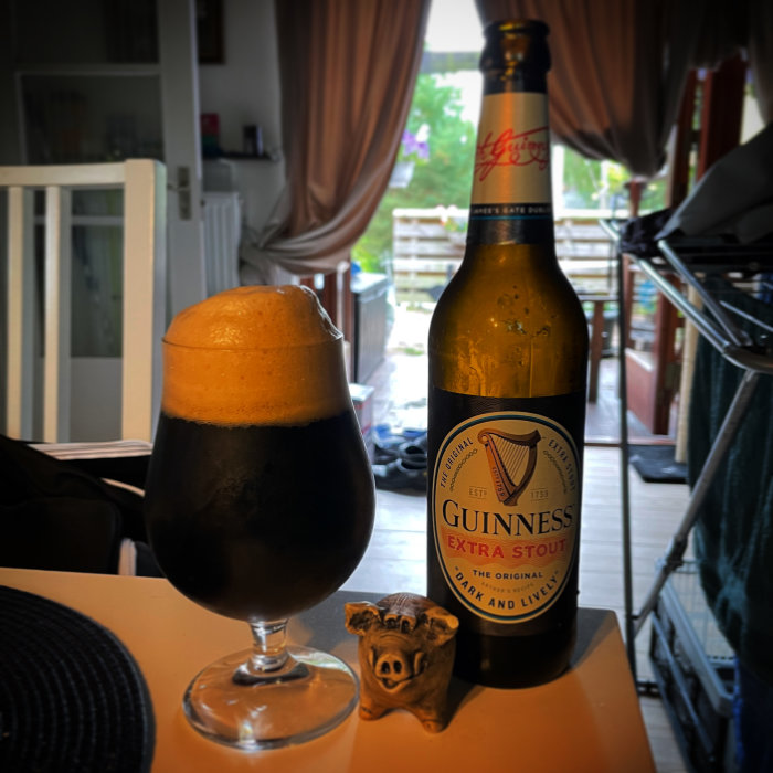 Ett glas med Guinness Stout med rikligt skum bredvid en flaska Guinness och en liten gristatuett.