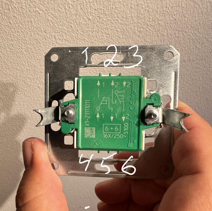 En hand håller en öppen elektrisk strömbrytare mot en vägg, numrerad för installation.