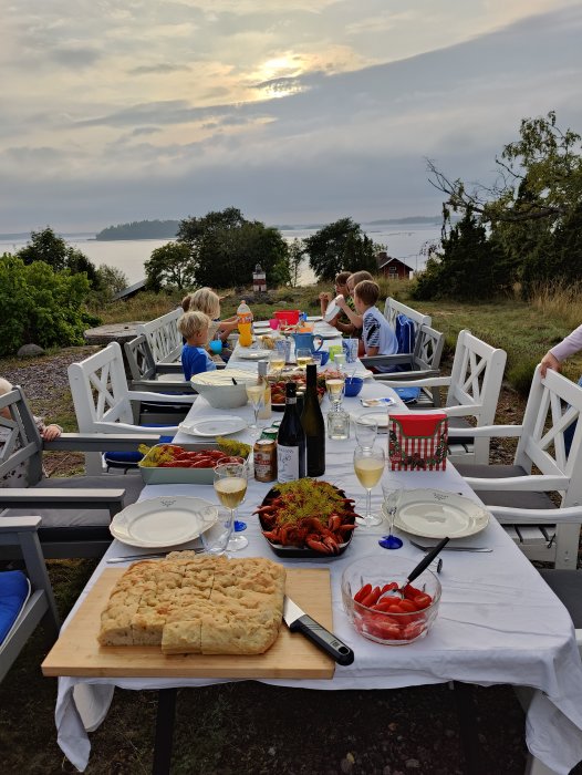 Utomhusbild på en dukad bord med kräftor, bröd och glas vin vid en kräftskiva med utsikt över vattnet.