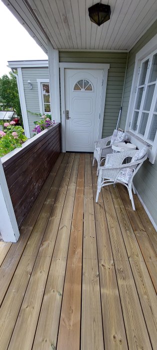 Inbyggd veranda med nytt trägolv, vit ytterdörr, vitmålad utemöbeluppsättning och blomlådor med rosor.