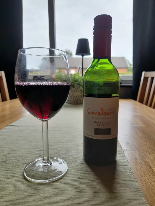 Ett glas rödvin och en flaska Malbec 2021 på ett bord framför ett fönster.