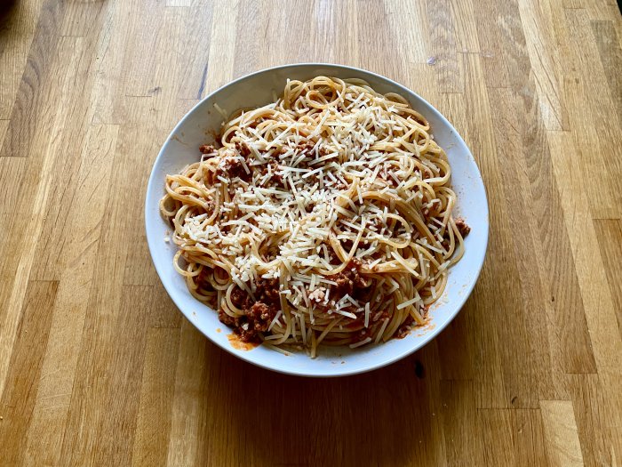 Stor portion spaghetti med köttfärssås och riven ost på träunderlag.