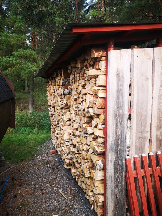 Vedstapel under tak bredvid rött staket i en trädgård.