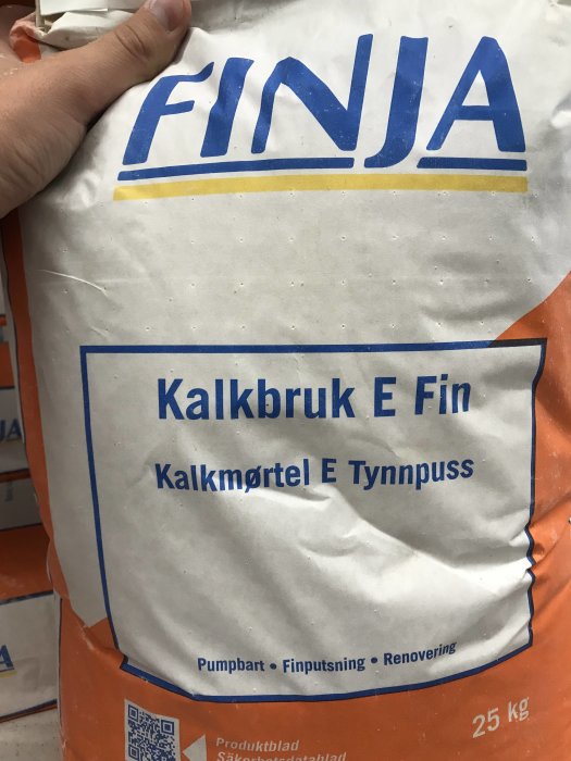 En hand håller en säck med FINJA Kalkbruk E Fin för finputsning och renovering på 25 kg.