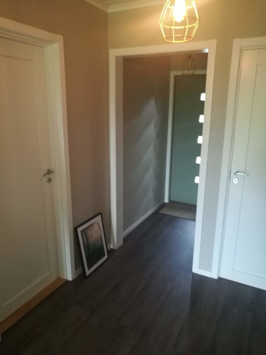 Korridor med grått laminatgolv, beigea väggar, vita dörrar och en tavelram lutar mot väggen.