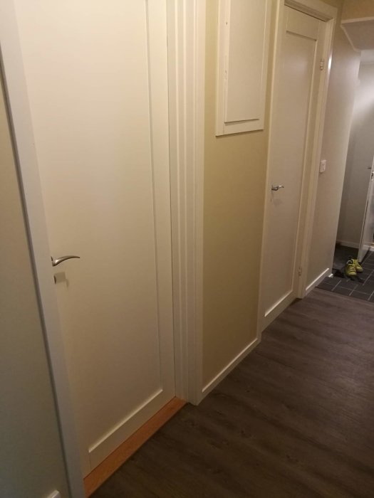 Ett ljust korridorsperspektiv med släta vitmålade dörrar och mörkt laminatgolv, detalj av skoskydd syns i bakgrunden.