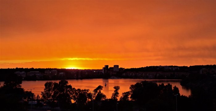 Solnedgång över stadssiluett med intensivt orange himmel och reflektioner i vatten.