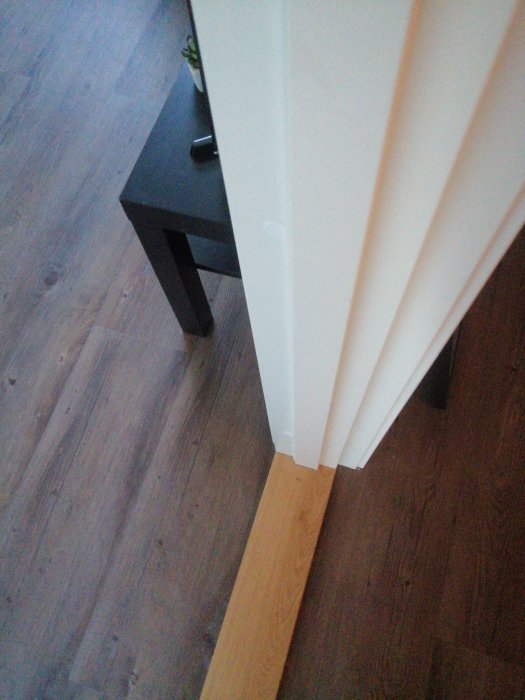 Vit dörr öppnas in till ett rum med mörkt sidobord och laminatgolv, synlig tröskel.